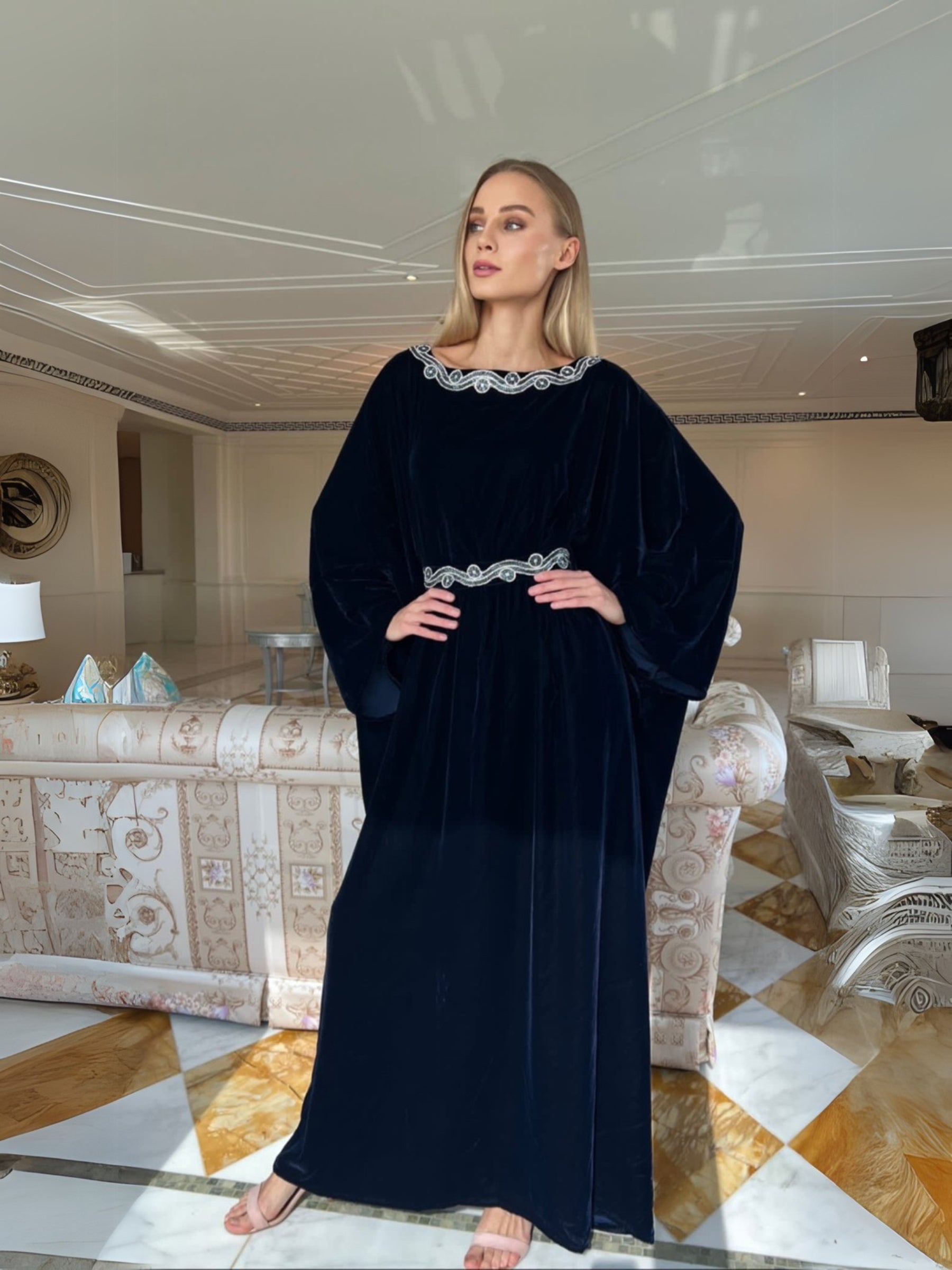 A woman standing in a luxurious interior wearing a blue velvet traditional dress with embroidered belt and neckline.